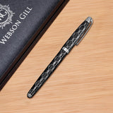 The WG ‘The Weaver’ Everyday Fountain Pen, featuring a silver lattice design and stainless steel medium nib, rests on a wooden surface next to its textured box, offering a smooth writing experience.