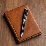 The WG 'The Boss' Everyday Fountain Pen, featuring a stainless steel nib and black finish, rests atop a closed brown leather notebook on a wooden surface.