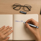 Someone writes "Webson Gill" in a grid notebook using WG’s ‘The Raider’ Everyday Fountain Pen, which has a black and silver design and a stainless steel medium nib; glasses lie nearby on the desk.
