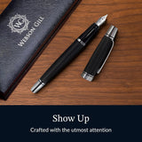 A black WG 'Show Up' Everyday Fountain Pen with a diamond guilloché pattern, cap off, rests beside its gift box on a wooden surface. Text: "Show Up.