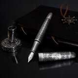 The Webson Gill Admiral Fountain Pen features an elegant antique silver design with intricate details, shown alongside a matching inkwell and a dark leather journal, all with an ornate silver finish.