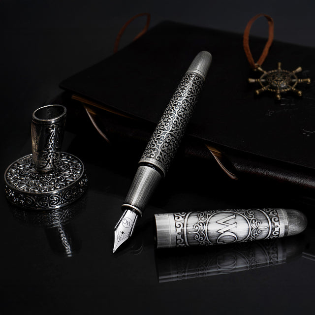 The Webson Gill Admiral Fountain Pen features an elegant antique silver design with intricate details, shown alongside a matching inkwell and a dark leather journal, all with an ornate silver finish.