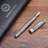 The Admiral Fountain Pen by Webson Gill, featuring an antique silver finish and elegant design with its cap off, is displayed beside a branded case on a wooden surface.