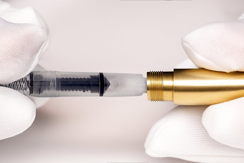 Gloved hands assembling a fountain pen, connecting a transparent ink converter to a gold barrel.