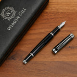 'The Mogul' Premium Fountain Pen by Webson Gill, featuring a stainless steel nib, black body, and silver accents, rests uncapped beside its Webson Gill case on a wooden surface.