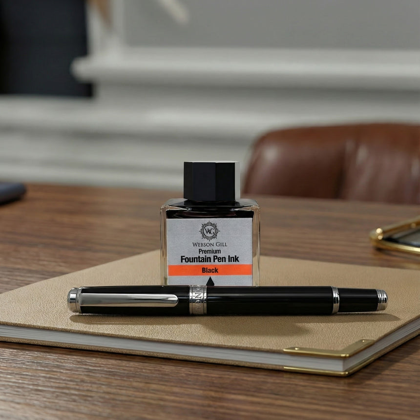 A bottle of black fountain pen ink and Webson Gill's 'The Mogul' Premium Fountain Pen with a stainless steel nib rest on a beige notebook atop a wooden desk.