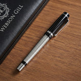 The WG ‘The Raider’ Everyday Fountain Pen, featuring a black and silver design with a stainless steel medium nib, rests on a wooden surface beside a book.