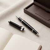 The Webson Gill 'The Mogul' Premium Ballpoint Pen, in black and silver with its cap, is displayed beside an open notebook and a brown case on a fabric surface. This executive pen delivers smooth ink flow, making it ideal for both professional and personal writing needs.