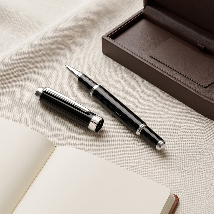 The Webson Gill 'The Mogul' Premium Ballpoint Pen, in black and silver with its cap, is displayed beside an open notebook and a brown case on a fabric surface. This executive pen delivers smooth ink flow, making it ideal for both professional and personal writing needs.