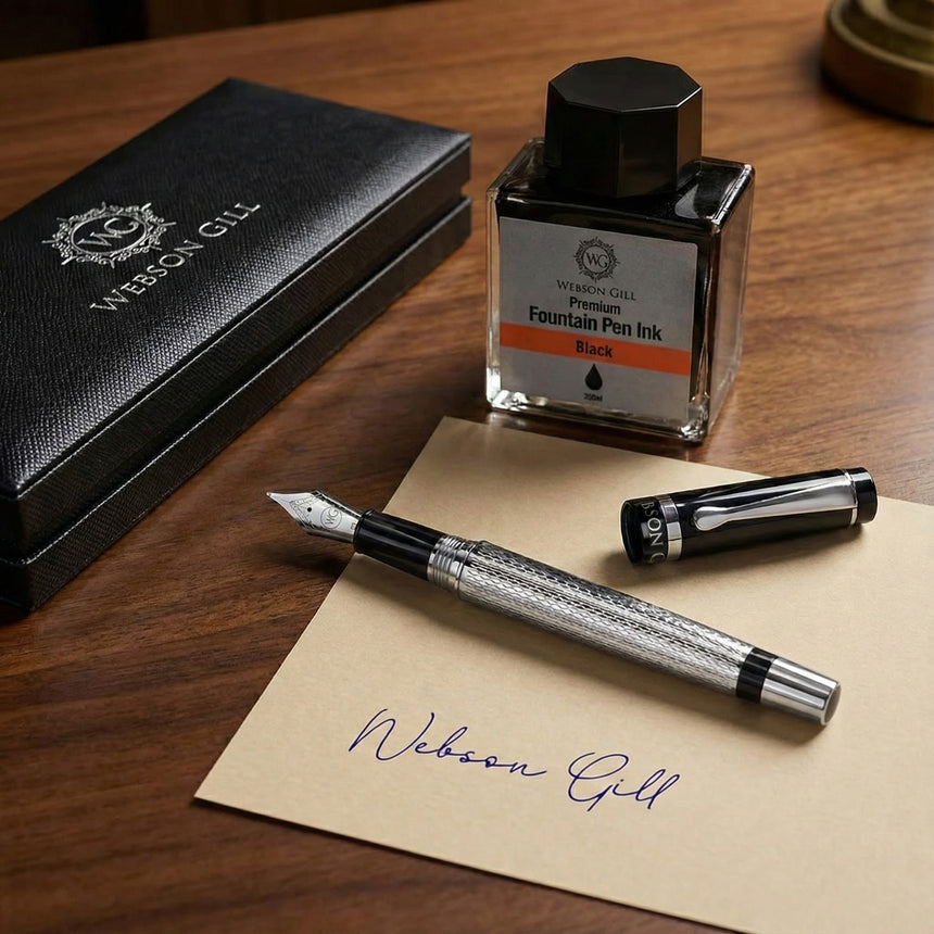 The WG ‘The Raider’ Everyday Fountain Pen, featuring a black and silver design with a stainless steel medium nib, rests by an ink bottle and black box on a desk next to an envelope labeled "Webson Gill" in blue ink.