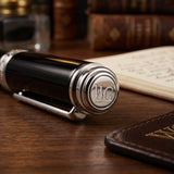 A black and silver 'The Mogul' Premium Ballpoint Pen by Webson Gill, engraved with WG and renowned for its smooth ink flow, rests on a wooden desk beside a notebook and books.