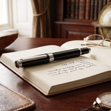 A Webson Gill "The Mogul" Premium Ballpoint Pen in black rests on an open notebook filled with handwritten notes, next to a pair of eyeglasses.