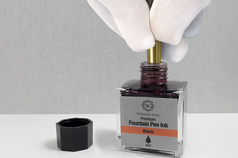 A gloved hand dips a pen into an open bottle of black fountain pen ink.