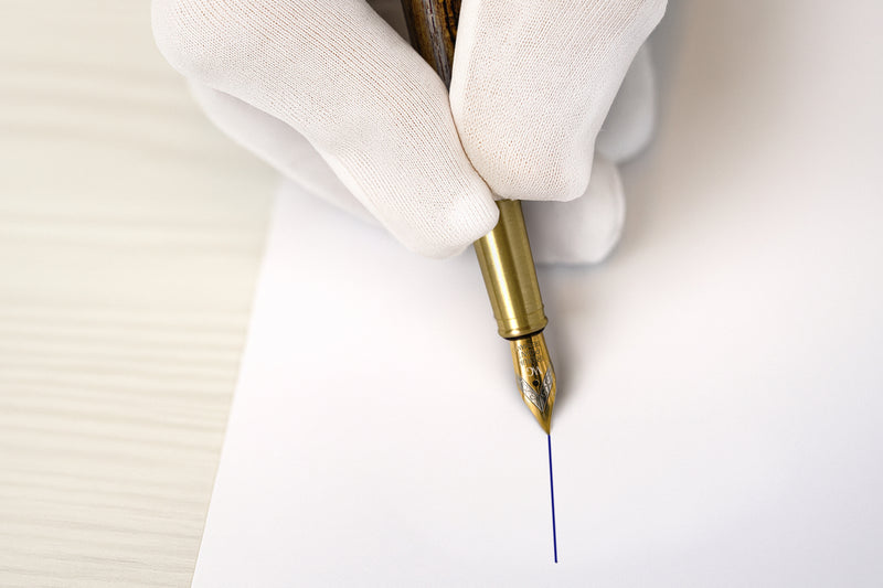 A gloved hand writes a straight blue line on white paper with a gold fountain pen.